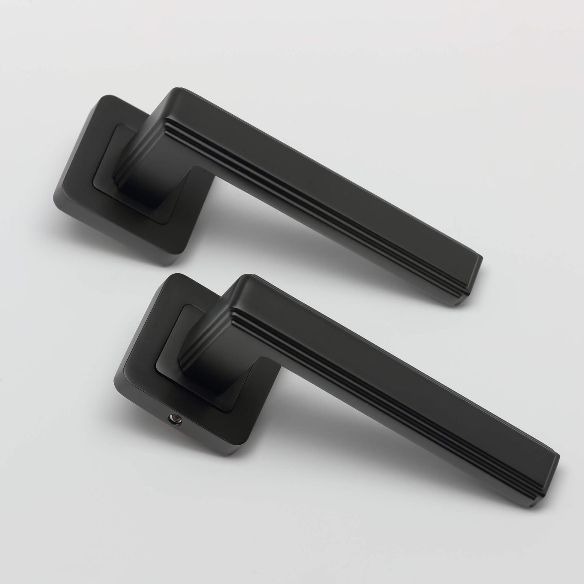 An angled view of a pair of Ares Flat Black Square Rose door handles on a grey background.