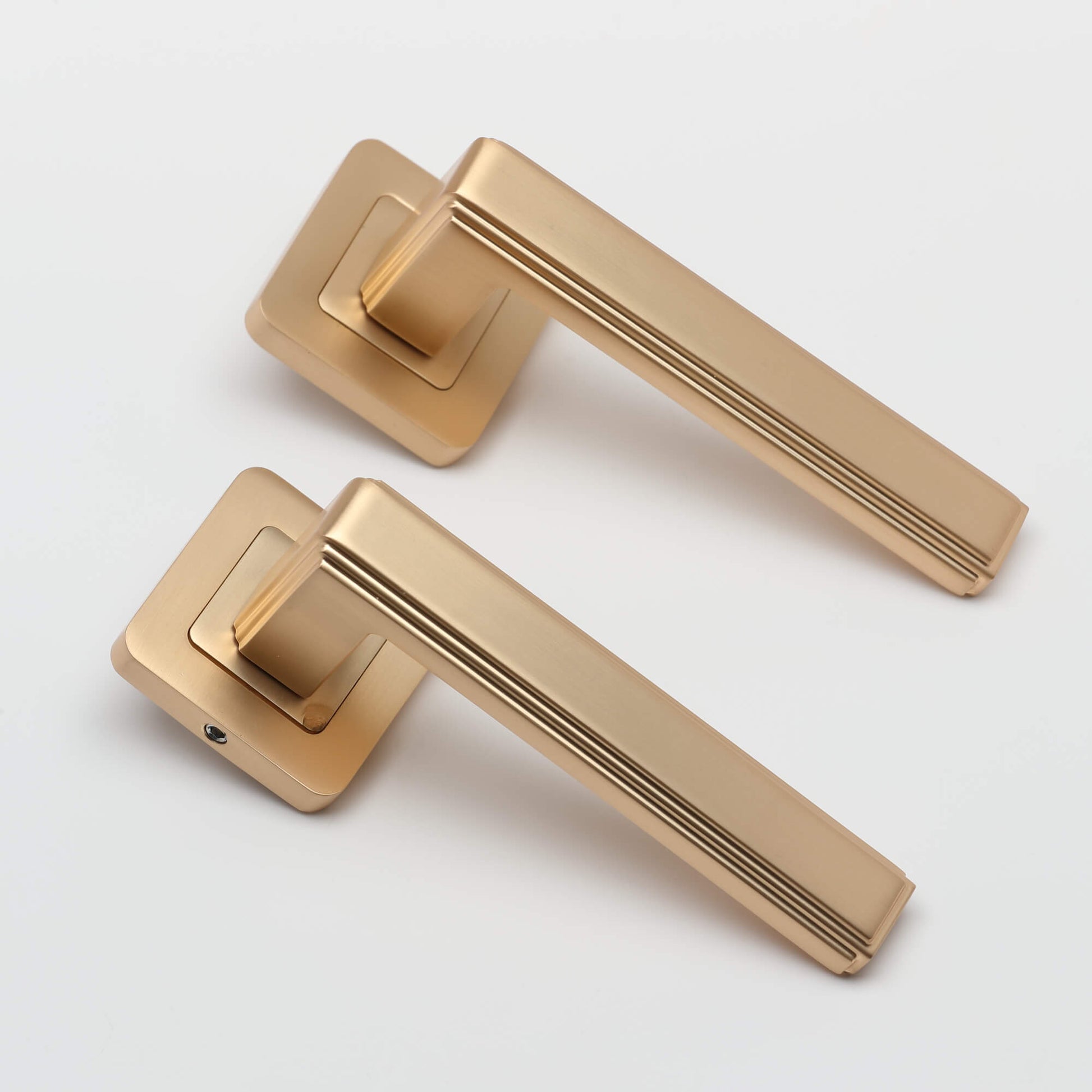 Angled view of a pair of Ares Satin Brass Square Rose door handles on a grey background.
