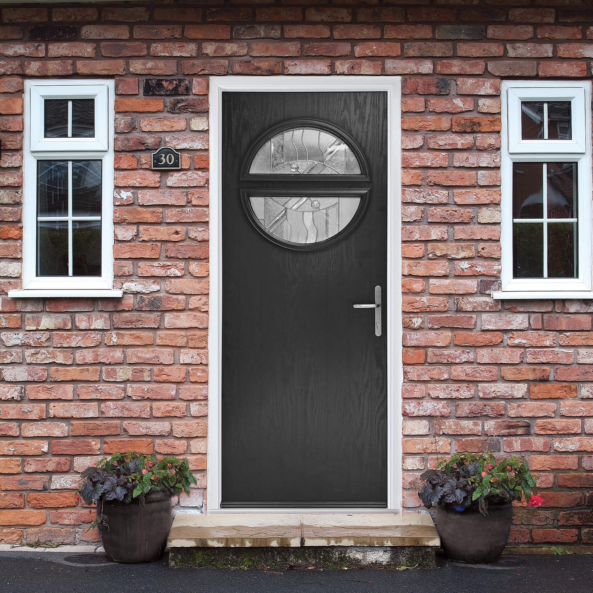 A Black Portsmouth Composite GRP External Door with round glass panel on a brick wall house exterior with two windows and potted plants.
